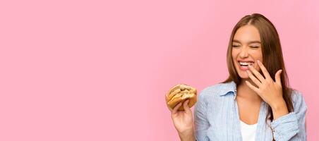 Fast Food. Happy Girl Eating Burger Laughing Posing On Yellow Studio Background. Panorama, Copy Space photo