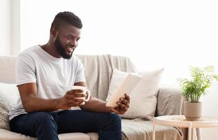 Black man sits on a couch in his home, relaxing with a cup of coffee and his tablet. He looks down at the screen, with a smile on his face. The image is a good representation of modern leisure photo