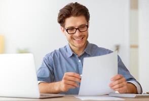 A man smiles as he reads a document while sitting at a desk with a laptop. photo