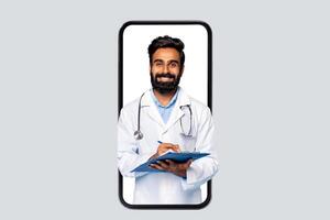 Indian man doctor is seen on the screen of a smartphone, wearing a white coat and stethoscope, smiling, and writing on a blue clipboard with a pen. photo