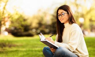 Asian woman is seated on the grass, focused on writing in a notebook. She holds a pen in her hand, with an open notebook on her lap. photo