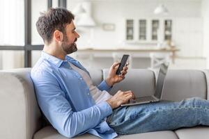 A man sits on a couch in a modern home, comfortably using a smartphone and a laptop. He is smiling, likely engaging with a digital task or enjoying some entertainment. photo