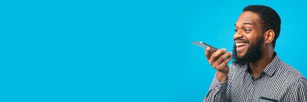 Happy young black man using voice assistant on smartphone, isolated over blue studio wall, copy space, web-banner photo