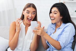 Happy Girl Showing Message On Phone To Friend Excited About Something Sitting On Sofa Indoor. Selective Focus photo