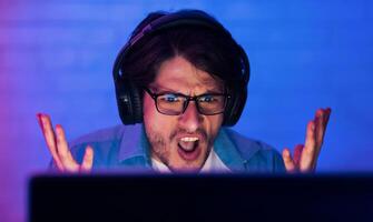 A young man wearing headphones looks frustrated while sitting in front of a computer screen in a room with blue lighting. He is looking directly at the camera with his mouth open in a scream. photo