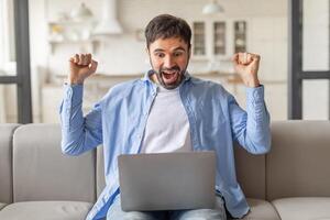A man sits on a couch, smiling widely and raising both fists in the air, while looking at a laptop screen. He appears to be celebrating a victory or success. photo