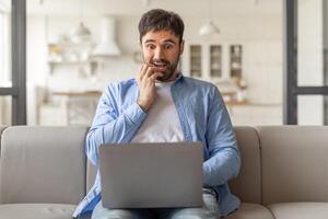 A man sits on a couch in his living room, biting his nails as he stares intently at a laptop in his lap. He looks concerned and apprehensive. photo
