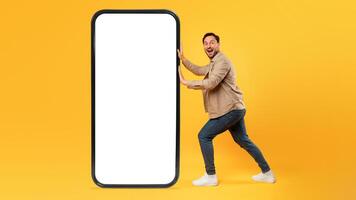 A cheerful man is standing next to a disproportionately large smartphone model with white blank screen, yellow background, mockup, copy space photo