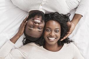Top view of romantic african american couple lying in bed together, tenderly touching each other and smiling at camera, closeup photo