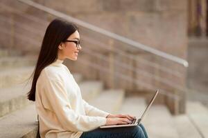 Asian woman is seated on a ledge, focused on her laptop computer. She appears concentrated and engaged in her work or task, with the cityscape visible in the background, blank space photo