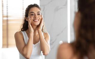 Perfecto piel. joven mujer conmovedor cara con hermosa suave y Fresco piel mirando en espejo en pie en moderno baño a hogar. hembra protección de la piel rutina. selectivo atención foto