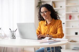 Happy Middle Eastern Businesswoman Using Laptop Typing And Working Online Sitting At Workplace Wearing Eyeglasses. Office Worker Lady Browsing Internet On Computer. Entrepreneurship. Side View photo