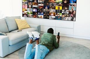 A man is lying on the floor of a living room, watching television. The television is displaying a grid of various images, and the man is holding a remote control in his hand. photo