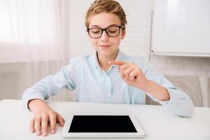 This image shows a young boy wearing glasses sitting at a white table. He is using a tablet and is pointing at the screen with his index finger. He appears to be focused on the device photo