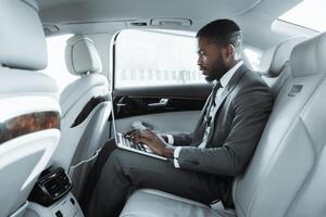 Black businessman in a suit sits in the back seat of a luxury car, working on a laptop computer. He is focused on his work, typing on the keyboard with his right hand photo