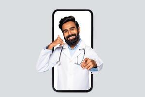 A smiling doctor in a white coat with a stethoscope is standing in a large smartphone. He is making a call me gesture with his left hand, and pointing at the viewer with his right. photo