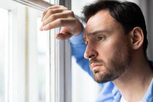 A man stands by a window, gazing out with a thoughtful expression. The natural light highlights his profile as he observes the view outside. photo