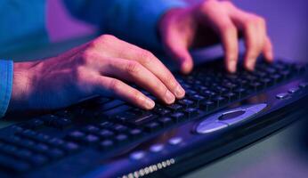 A close up image of a persons hands typing on a black computer keyboard. The keyboard has a built-in mouse pad. The image is lit by purple and blue light, giving it a futuristic or techy feel. photo