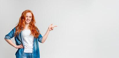 A cheerful red-haired woman stands against a plain background, one hand on her hip and the other extended as she points to the vacant area beside her, copy space photo