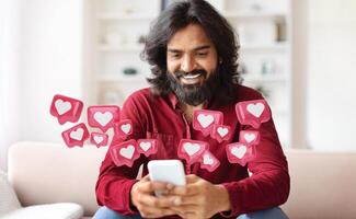 Hindu man with a long beard is sitting on a couch and smiling while looking at his smartphone. There are many heart icons surrounding him, suggesting he is receiving lots of likes photo