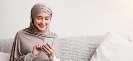 contento árabe hembra en hijab desplazamiento social redes o acecho foto en teléfono inteligente a hogar, sentado en cómodo sofá, gratis espacio