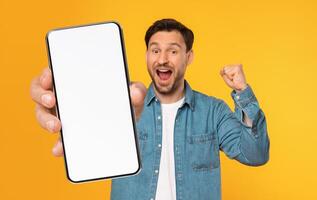 A cheerful man with a denim jacket joyfully raises a smartphone with a blank screen, showcasing an expression of excitement. The bright yellow backdrop adds to the lively atmosphere. photo