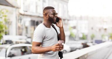 A man with a beard and sunglasses walks down a city street while talking on his phone and holding a to-go cup of coffee. photo