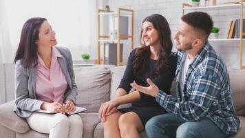 A young couple is sitting on a couch in an office setting, talking with their therapist. The therapist is a woman, and she is listening intently to the couple photo