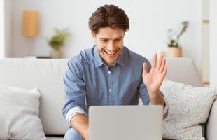 A man sits on a couch, smiling and waving to the laptop screen in front of him. photo