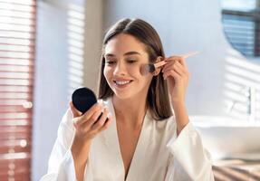 Daily Makeup. Smiling Young Female Applying Blush With Makeup Brush In Bathroom, Beautiful Happy Woman Wearing White Silk Robe Using New Cosmetics While Getting Ready At Home, Copy Space photo