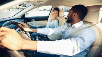 African American man with a beard is driving a car while a woman sits in the passenger seat. He is looking forward, and she is smiling at him. The car is in motion, and they are in a city environment. photo
