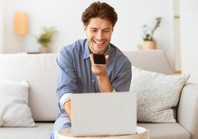 A man is sitting on a couch, smiling while looking at a smartphone in his hand. He appears to be using a voice assistant to complete a task while working on a laptop. photo