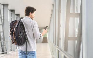 Booking Hotel Online. Smiling Man Holding Backpack And Using Smartphone In Airport Terminal Corridor, Rear View With Free Space photo