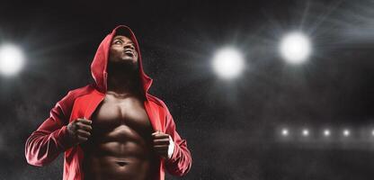 A muscular athlete in a red hoodie stands with his chest exposed, looking up at the stadium lights. photo