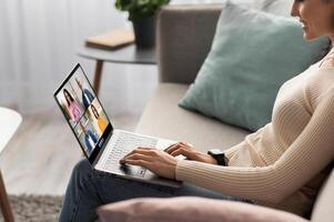 A woman sits on a couch at home and uses a laptop computer to participate in an online conference meeting. She is smiling and appears engaged in the conversation, side view photo
