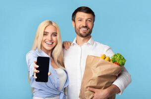 A smiling woman holds a smartphone with a black screen, while a man next to her holds a paper bag full of groceries. They are both looking directly at the camera and smiling. photo