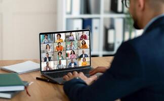 A businessman sits at a desk with a laptop open in front of him, showing a conference screen with many people in individual squares. He is using his laptop for an online meeting. photo