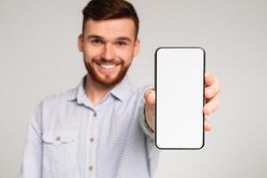 Young guy showing his phone with blank space on camera and smiling, isolated on grey studio background, mockup photo
