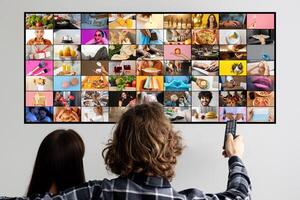A couple sits on a couch, facing a television screen filled with a collage of images. The woman sits with her back to the camera while the man holds a remote control and faces the screen photo
