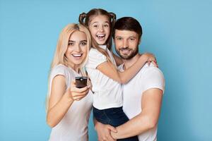 A happy family is watching television together at home. The parents are smiling and the daughter is holding onto her father while watching the screen. photo