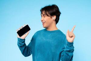 Asian young guy in a blue sweater is happily listening to music on wireless earbuds while holding his smartphone. He points upwards with a smile on his face photo