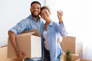 Joyful Afro Spouses Showing Key Embracing And Holding Moving Box After Relocation Standing In New Flat. Selective Focus photo