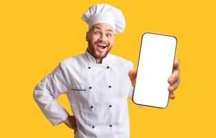Food Delivery App. Excited Chef Guy Showing Mobile Phone Empty Screen Posing On Yellow Background. Studio Shot photo