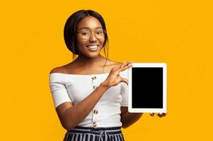 Afro Young Lady Showing Digital Tablet Empty Screen Smiling At Camera Standing On Orange Background In Studio. Mockup photo