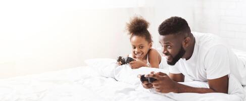 African American father and his daughter are lying in bed, both holding game controllers and smiling. They are both wearing white shirts and are looking at the camera, copy space photo