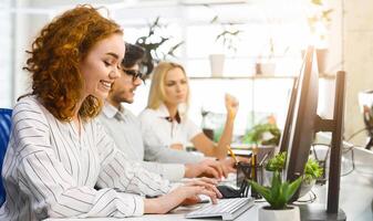 Several coworkers are seated at a desk, each engrossed in their computer screens, actively typing and clicking. The group appears focused and concentrated on their tasks photo