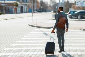 African American businessman in a suit crossing the street with a rolling suitcase, seen from behind in an urban setting, back view photo