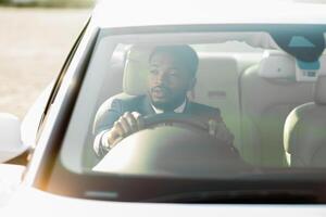 Black man wearing a suit sits in the drivers seat of a car. The car is facing towards the sun, creating a bright, hazy effect. He is gripping the steering wheel and looking straight ahead photo