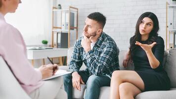 A couple sits on a couch in an office, talking to a therapist. The woman is talking and gesturing with her hand, while the man is listening and thinking thoughtfully. The therapist is taking notes. photo