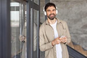 A man wearing headphones and a beige button-up shirt is leaning against a glass door and using his smartphone. He is smiling and appears to be enjoying listening to music or audio content. photo